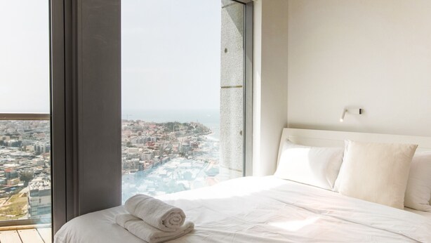 A bedroom with a large window offering a city view, a neatly made bed, and a white headboard. - MIKLAT Luxury Condo & Spectacular View by FeelHome (Tel Aviv - Jaffa)