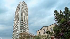 A tall, modern high-rise building with balconies, surrounded by greenery and a historic building. - MIKLAT Luxury Condo & Spectacular View by FeelHome (Tel Aviv - Jaffa)