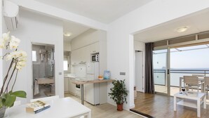 Eine moderne Küche mit weißen Schränken, einer Mikrowelle und einem Kühlschrank. Es gibt einen Essbereich mit einem weißen Tisch und Stühlen sowie einen Balkon mit Meerblick.