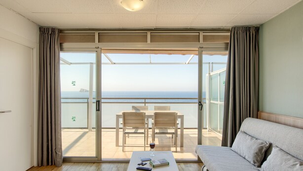 Ein Zimmer mit Balkon, einer Couch, einem Tisch mit einem Weinglas und Meerblick.