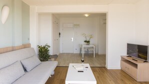 Ein modernes Wohnzimmer mit einer Couch, einem Couchtisch mit Buch und Glas, einem Fernseher auf einem Holzboden und einem Essbereich im Hintergrund.