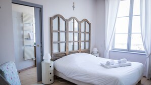 Un dormitorio con cama, una silla, una mesita de noche, una lámpara y una ventana con cortinas.