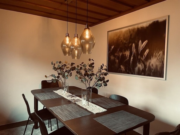 Un comedor con mesa de madera, sillas y lámparas colgantes. En la pared, sobre la mesa, hay una obra de arte enmarcada.