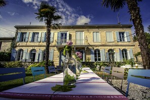 A table set with a vase of flowers and a bottle, positioned in front of a building with blue shutters and a balcony.