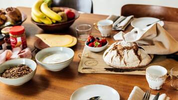 Une table de salle à manger en bois, sur laquelle on trouve un pain, un bol de fraises, une assiette de fromage en tranches et un pot de beurre de cacahuète.