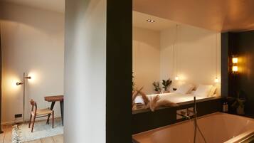 A modern bathroom with a large bathtub, a mirror, and a bedside table with a chair.