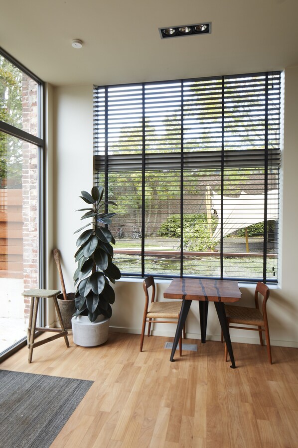 A room with a large window, a wooden table, two chairs, a potted plant, and a small stool. - Chambre d'Amis by Alix (Ghent)