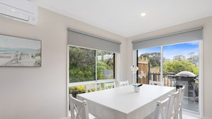 A modern dining area with a white table and chairs, large windows with a view of greenery and a barbecue area. - This revamped quintessential holiday home, prime location (sleeps 8) (Sorrento)