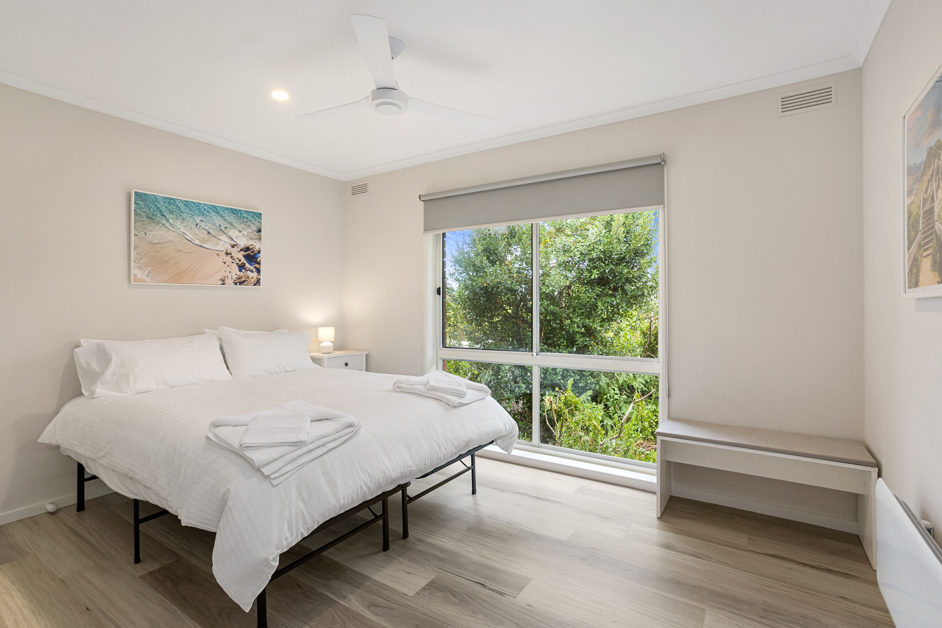 A bedroom with a bed, a nightstand, a ceiling fan, a window with a view of greenery, and a painting on the wall.