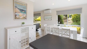 A modern kitchen with white cabinetry, a dark countertop, and a dining area with white chairs and a table. There is a large window providing a view of greenery outside. - This revamped quintessential holiday home, prime location (sleeps 8) (Sorrento)