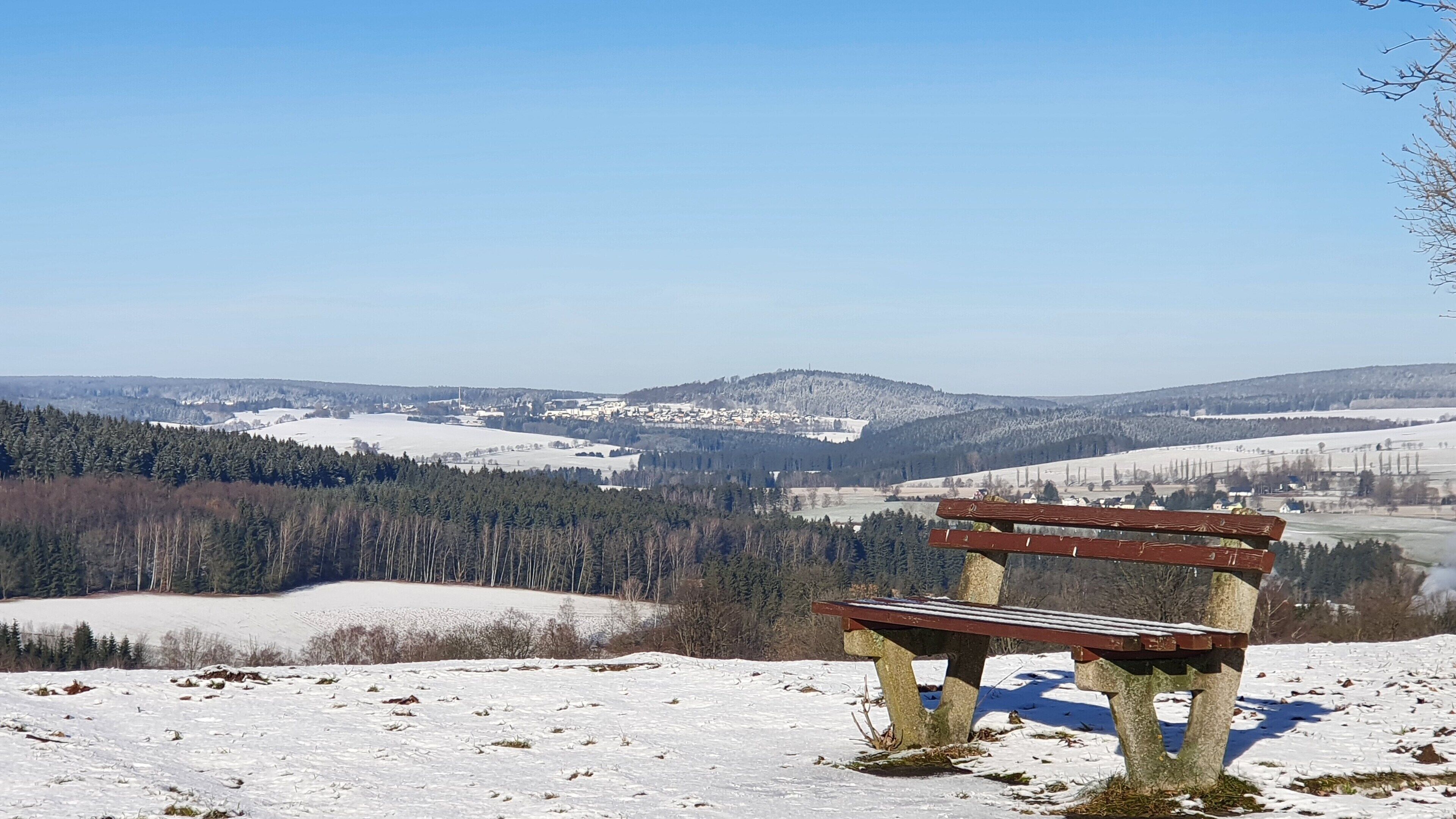 A bench on a snow-covered hill overlooking a town with buildings and trees.