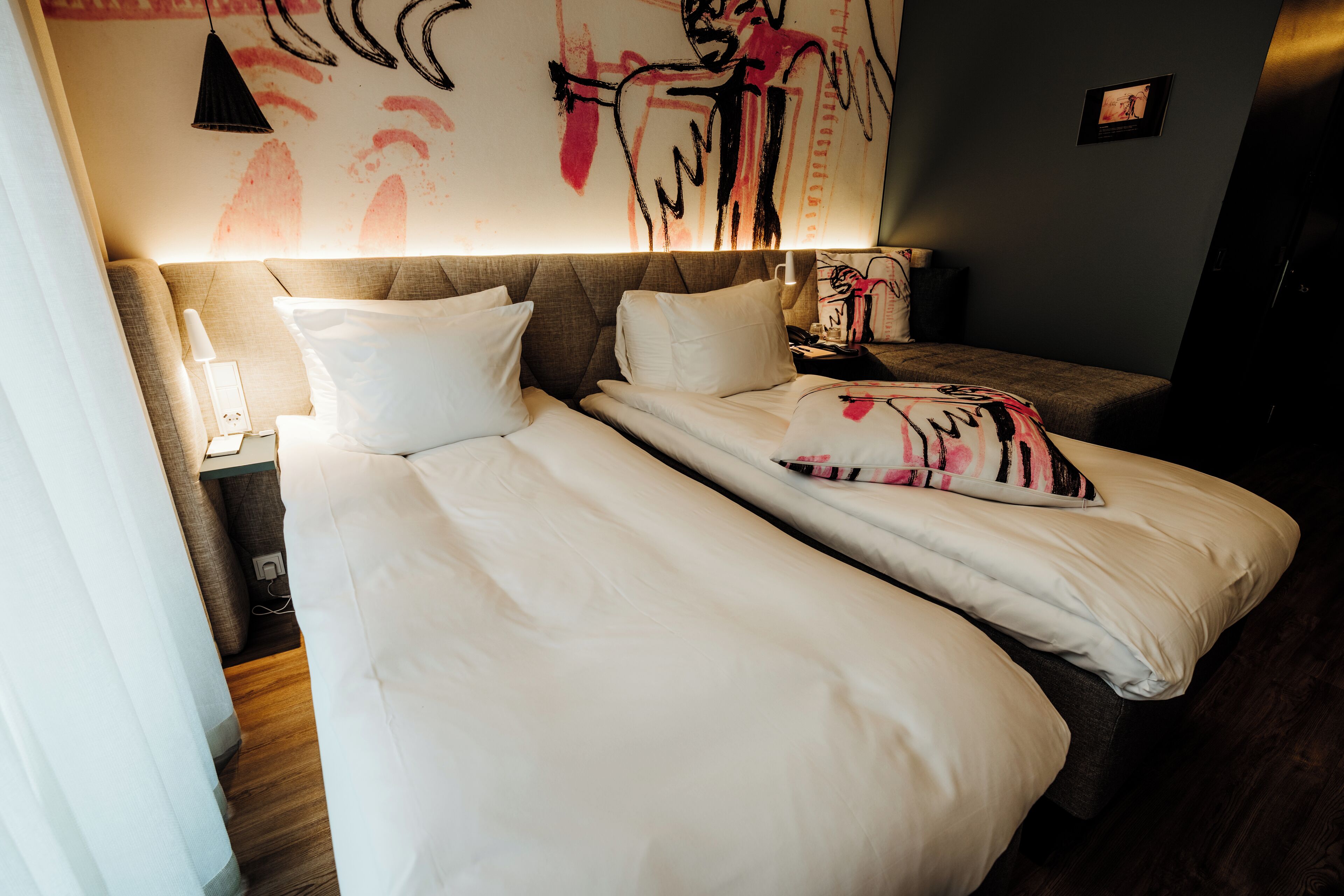 A double bed with white bedding and a striped pillow, a bedside table with a lamp, and a wall with abstract art.