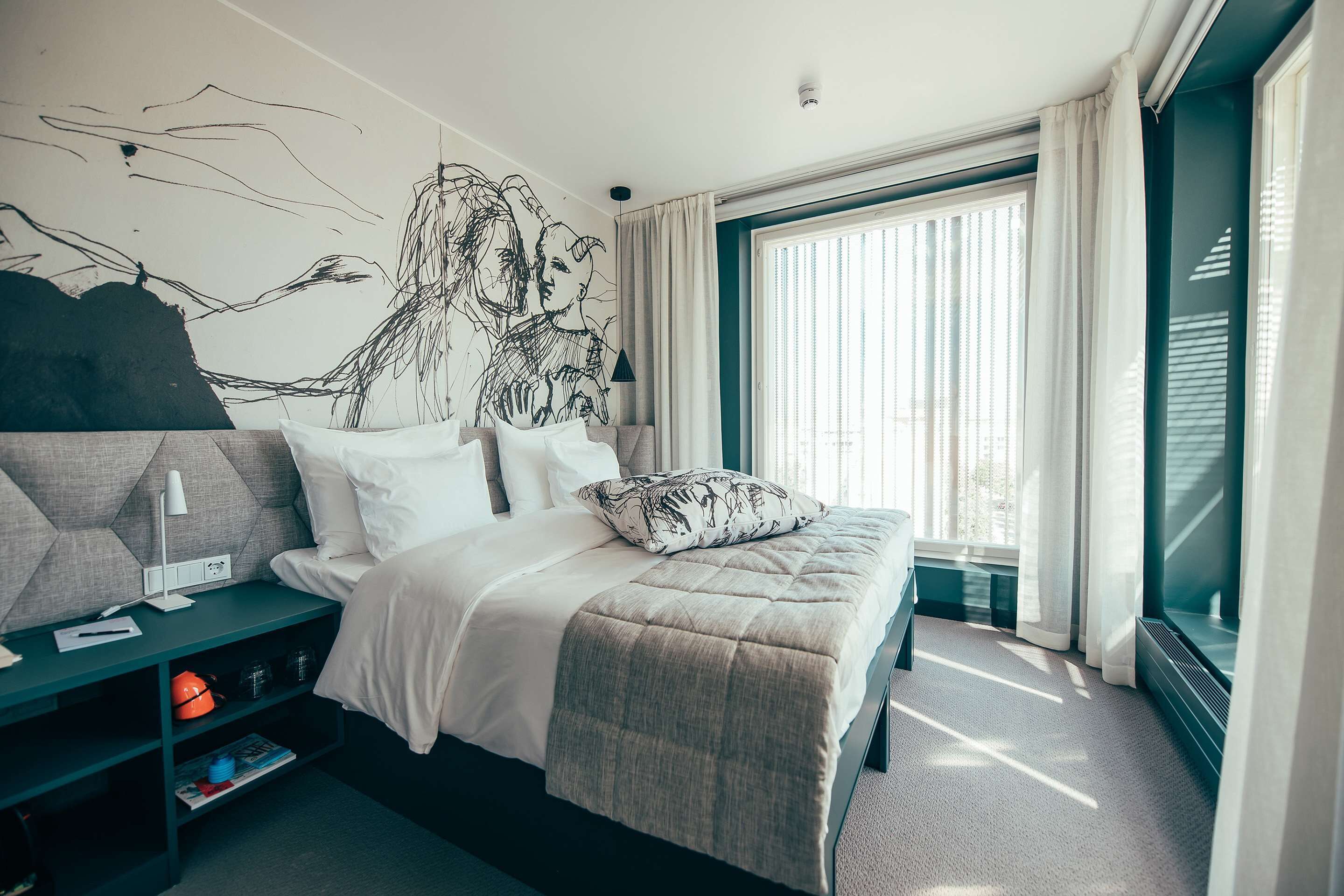 A modern hotel room with a large bed, a bedside table with a lamp, a wall mural, and a large window with curtains.