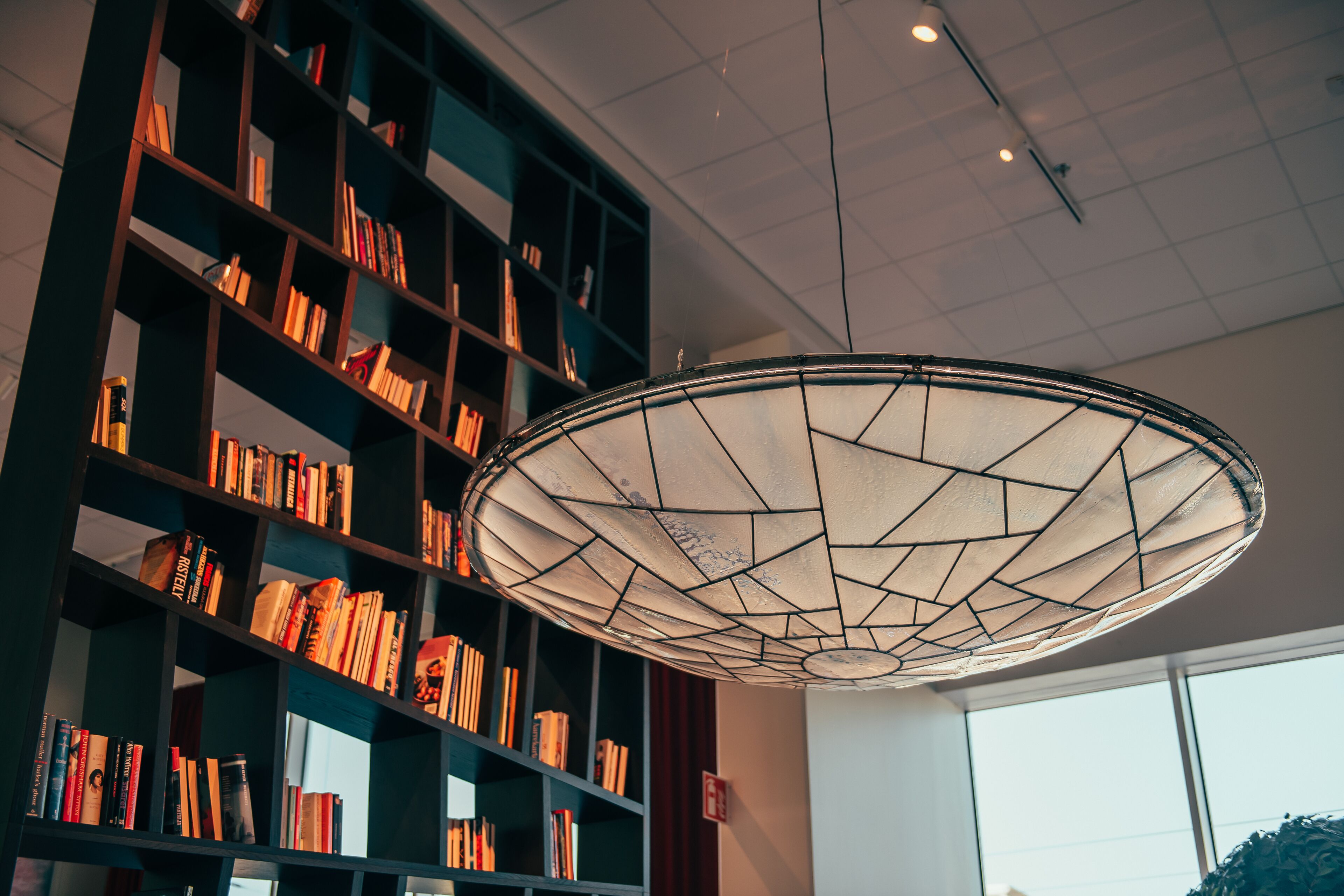 A modern interior with a large bookshelf filled with books, a geometric ceiling light fixture, and large windows allowing natural light.