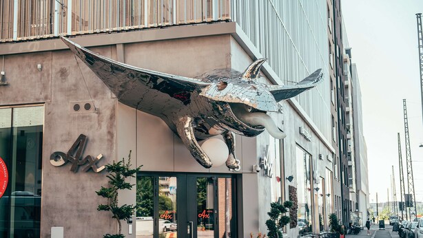 Ein Gebäude mit einer großen metallischen Skulptur eines Stiers, inklusive Kopf und Hörner, einer Fassade mit der Aufschrift "Ax" und Außenplätzen mit Pflanzen in Töpfen.