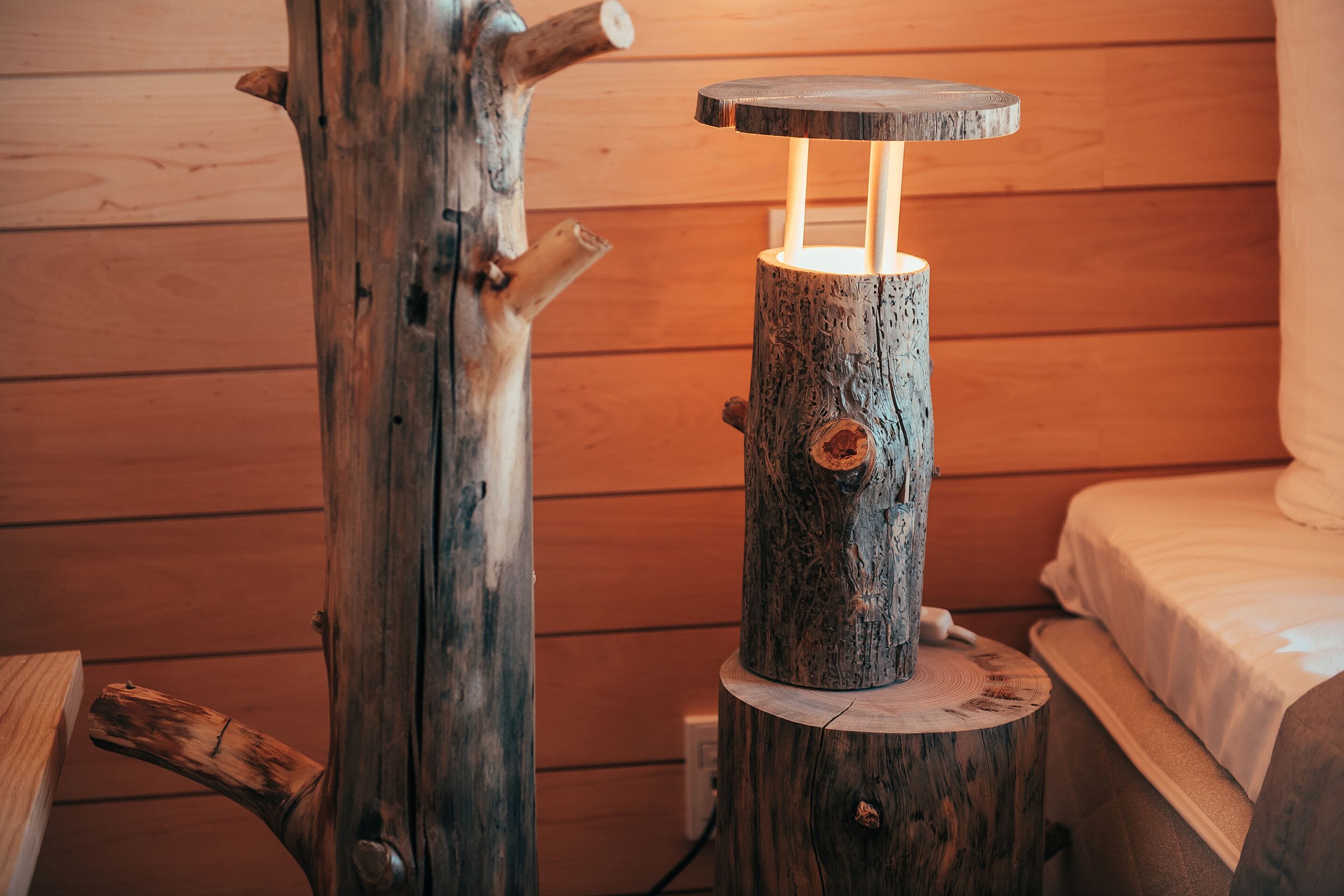 Una mesa de madera rústica con una fuente de luz integrada, colocada junto a una cama en una habitación de madera.