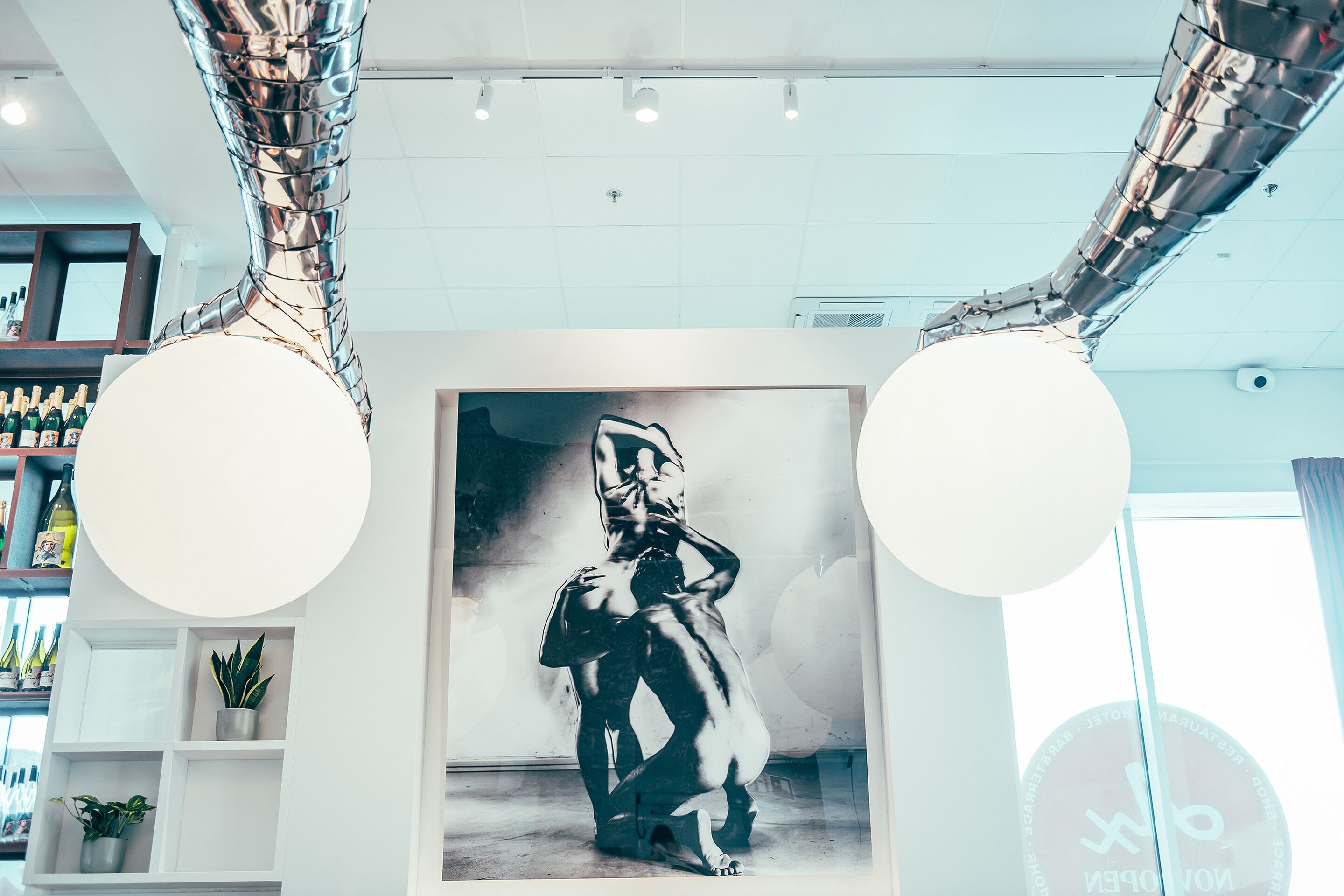 A modern interior with a large framed artwork of a human figure, metallic structural elements, and a bar area with bottles.