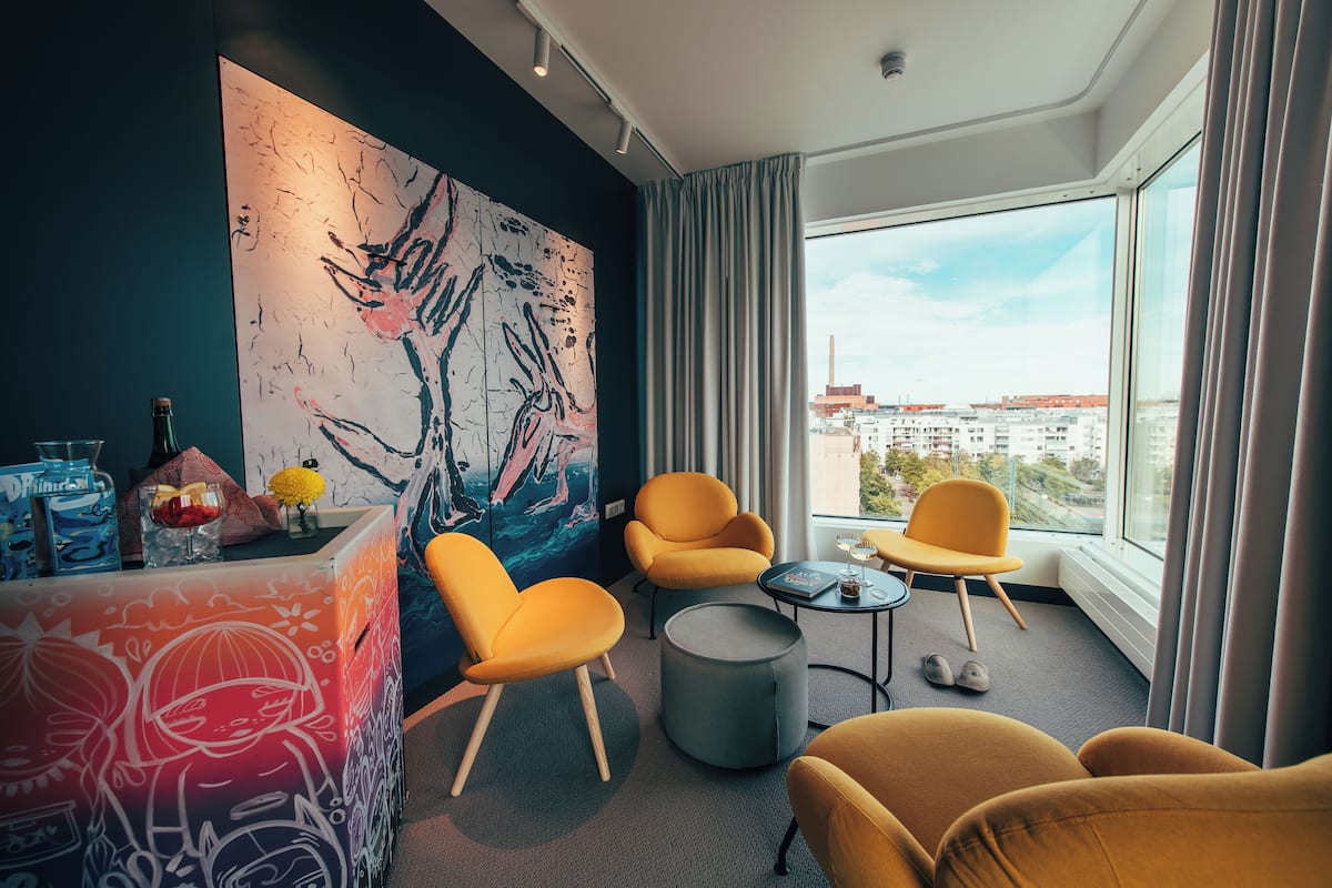 A modern hotel room with a large abstract painting, comfortable seating, a small table, and a view of the city through large windows.