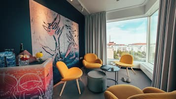 A modern hotel room with a large abstract painting, comfortable seating, a small table, and a view of the city through large windows.