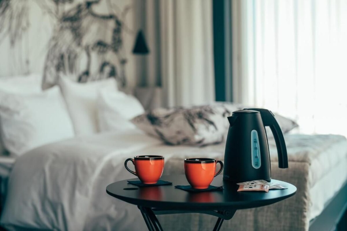 A neatly made bed with white linens and a pillow, a small round table with two orange cups and a black kettle, and a window with curtains in the background.