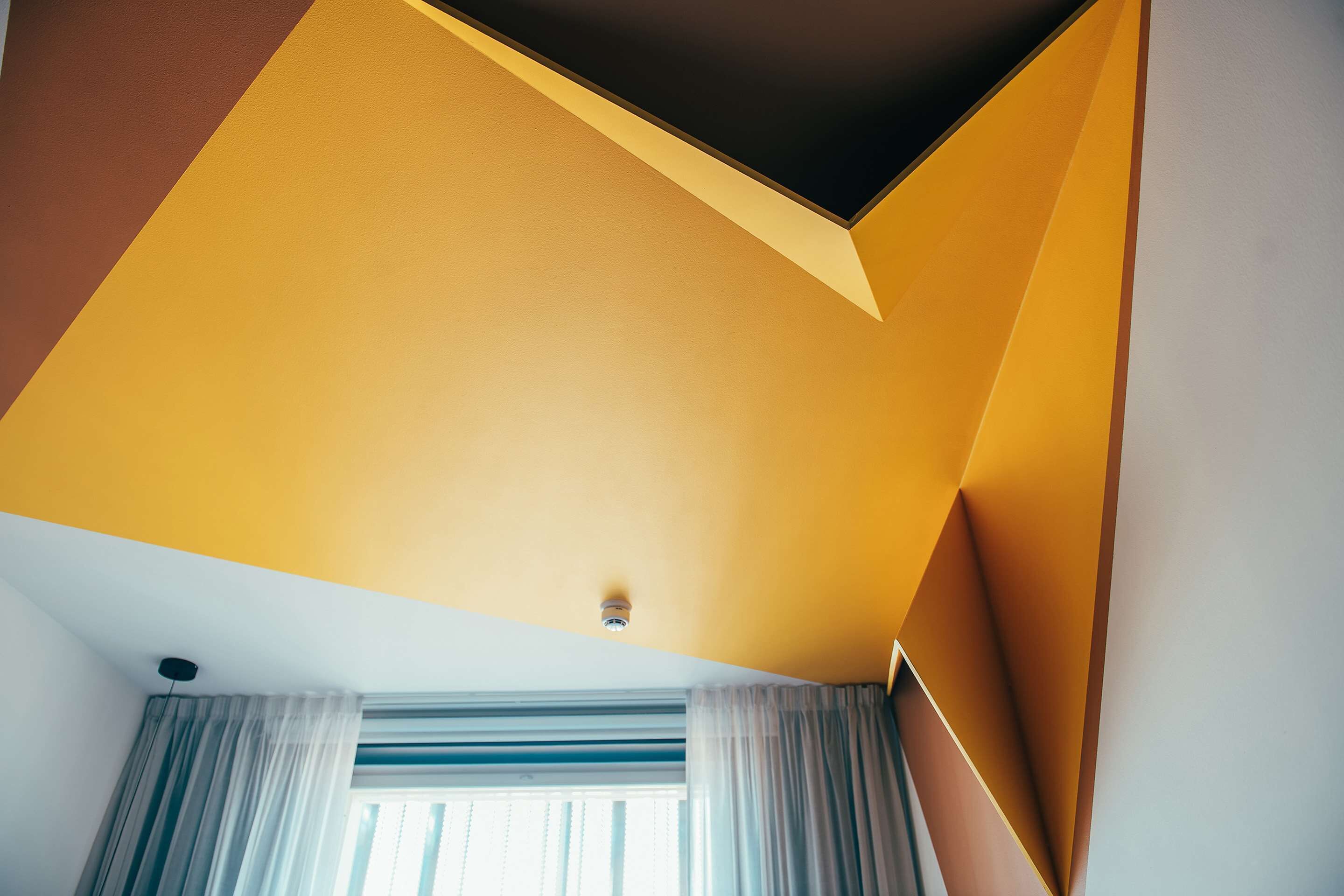 Une pièce avec un plafond en pente, une fenêtre avec des rideaux et un luminaire apparent.