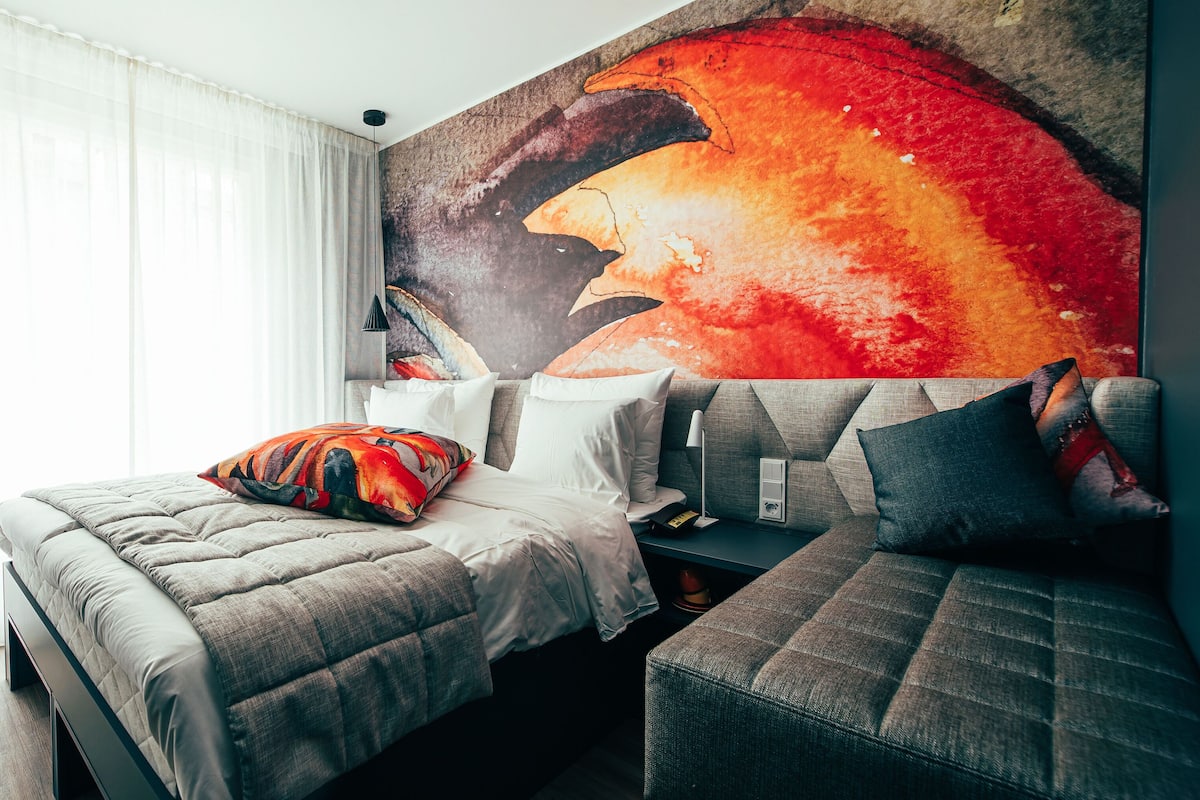 A hotel room with a bed, a sofa, a large wall mural, and a window with sheer curtains.