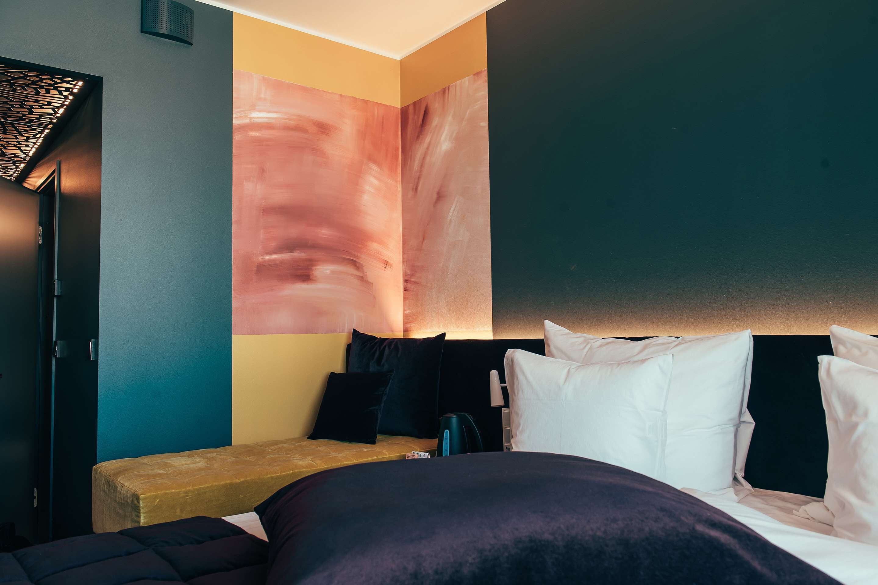 A modern hotel room with a bed, a bench, and a large abstract painting on the wall.