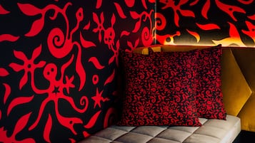 Una cama con un cojín estampado, una pared con diseños abstractos en rojo y una lámpara con forma geométrica.