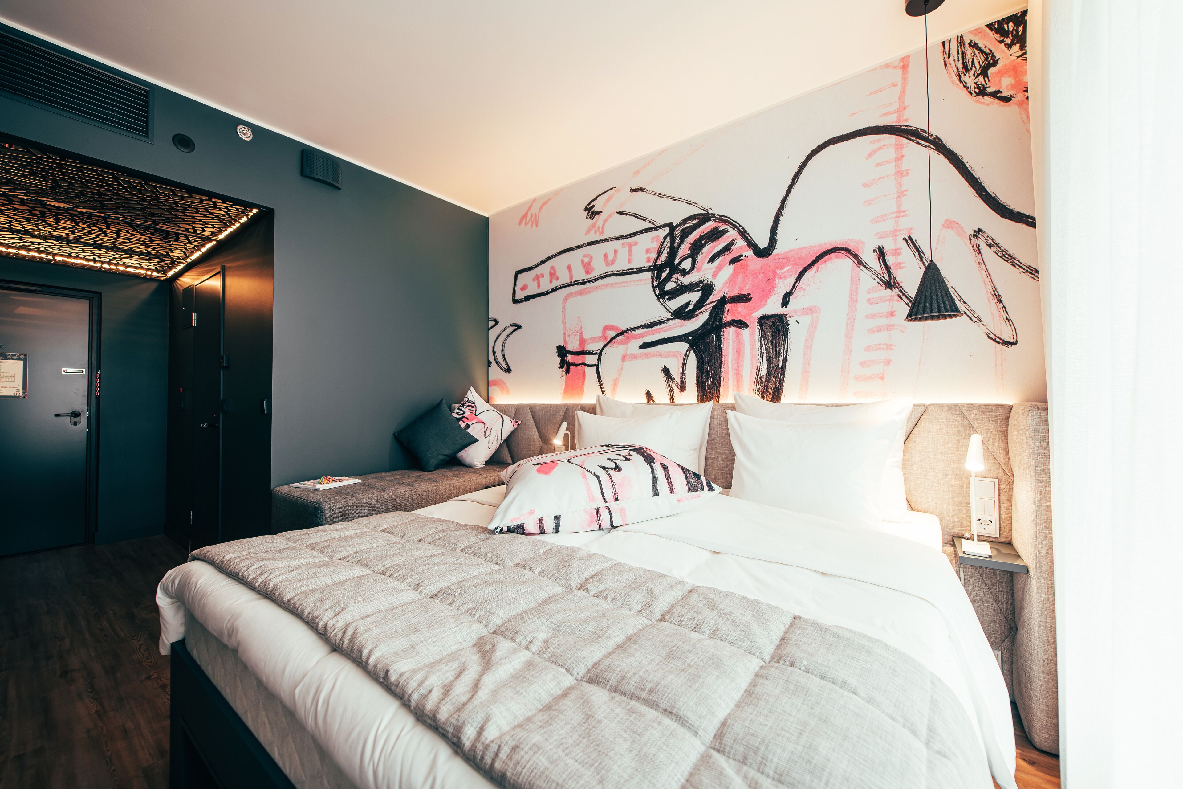 Una habitación de hotel con cama, mesita de noche, lámpara, una obra de arte en la pared y una puerta.