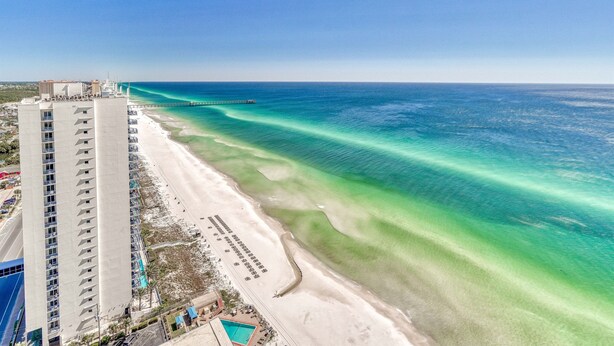 A high-rise hotel adjacent to a beach with clear turquoise waters and a pier extending into the sea.