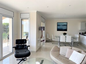 A modern living room with a glass coffee table, a beige sofa with cushions, a black office chair, a dining area with a glass table and chairs, and a large window offering a view of the outdoors.