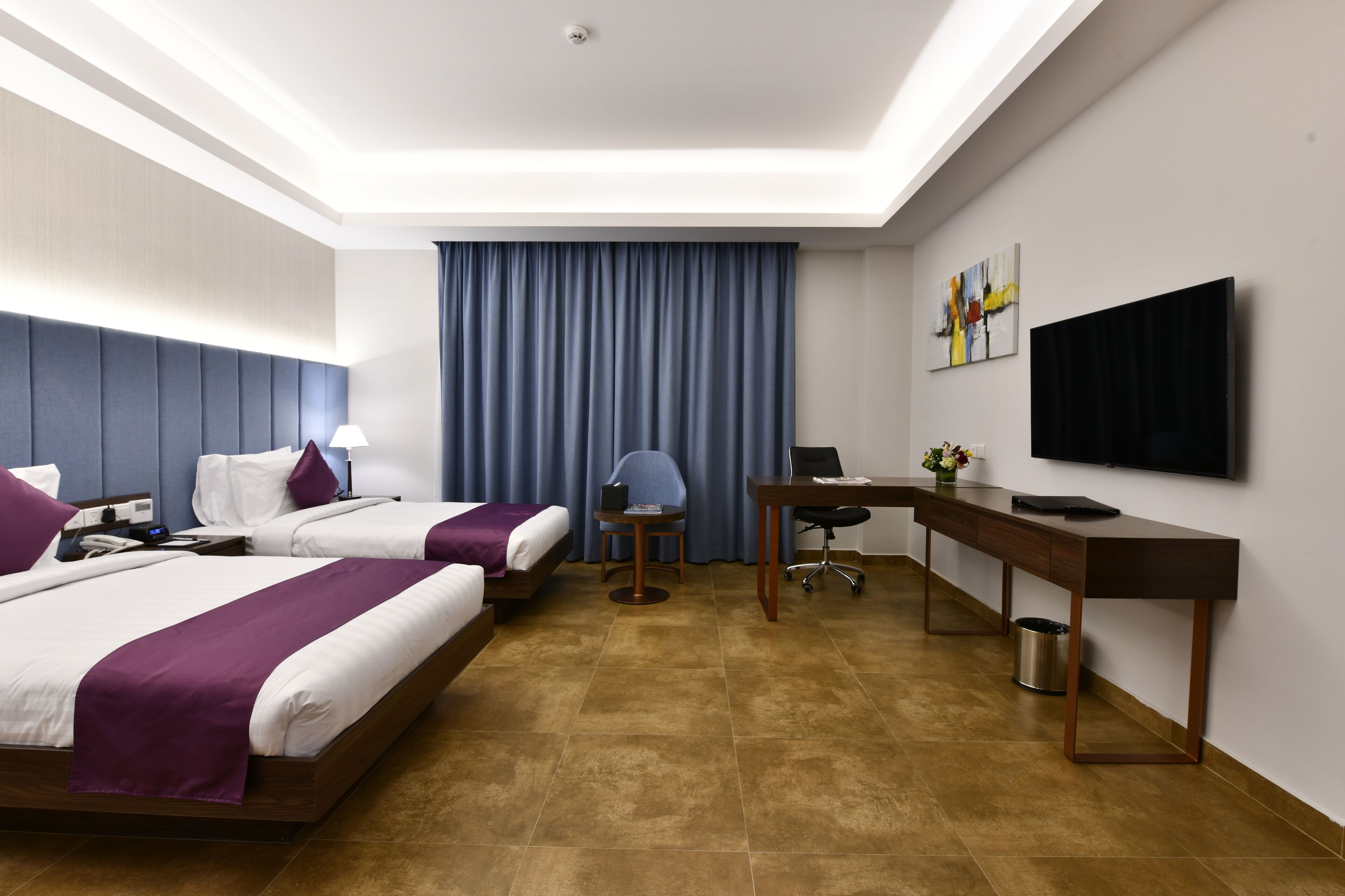 Habitación de hotel con dos camas, un escritorio, una silla, un televisor y un ventanal con cortinas.