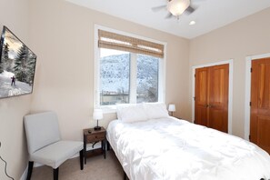 Quarto com cama, cadeira, mesa de cabeceira, janela com vista para colinas cobertas de neve e duas portas de madeira.