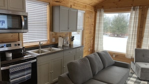 A cozy cabin interior with a kitchenette, grey sofa, and a view of a snowy landscape. - Tiny house, loft bedroom, near the river, Cooperstown and Baseball hall of fame (Richfield Springs)