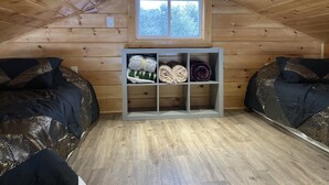 A cozy attic bedroom with wooden walls and a sloped ceiling. There are two beds with patterned bedding, a window with a view of trees, and a shelf with rolled blankets. - Tiny house, loft bedroom, near the river, Cooperstown and Baseball hall of fame (Richfield Springs)