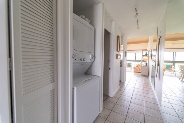 Un cuarto de lavado con lavadora y secadora, piso de baldosas y vista al área de estar a través de una puerta abierta.