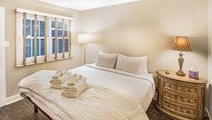 A neatly made bed with a white comforter and pillows, a bedside lamp, a wooden dresser with drawers, and a window with blinds. - Pet friendly, Historic Garden Level w/ King Bed & No Stairs by Lucky Savannah (Savannah)