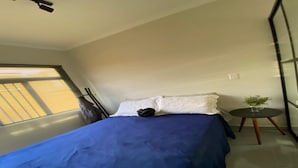 A bedroom with a bed covered in a blue bedspread, a white pillow, and a small round table with a plant. - The building is right across from the beach, and the studio apartment has a wide view of it. (SP)