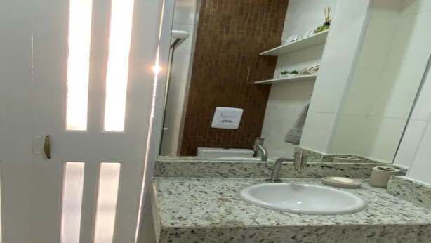 A bathroom with a granite countertop, a white sink, and a mirror. There is a wooden door with vertical slats and a white wall with a small shelf. - The building is right across from the beach, and the studio apartment has a wide view of it. (SP)