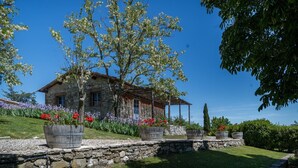 A stone house with a wooden porch, surrounded by a garden with various flowers and a clear blue sky.