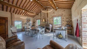 A rustic kitchen with a brick wall, wooden beams, and a large oven. There is a dining area with metal chairs and a table, and a wooden bench in the foreground.