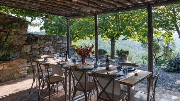 An outdoor dining area with stone walls, wooden tables, and chairs, surrounded by greenery and a view of the landscape.