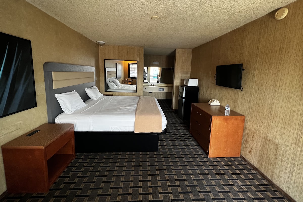 Quarto de hotel com cama, escrivaninha, televisão e espelho.