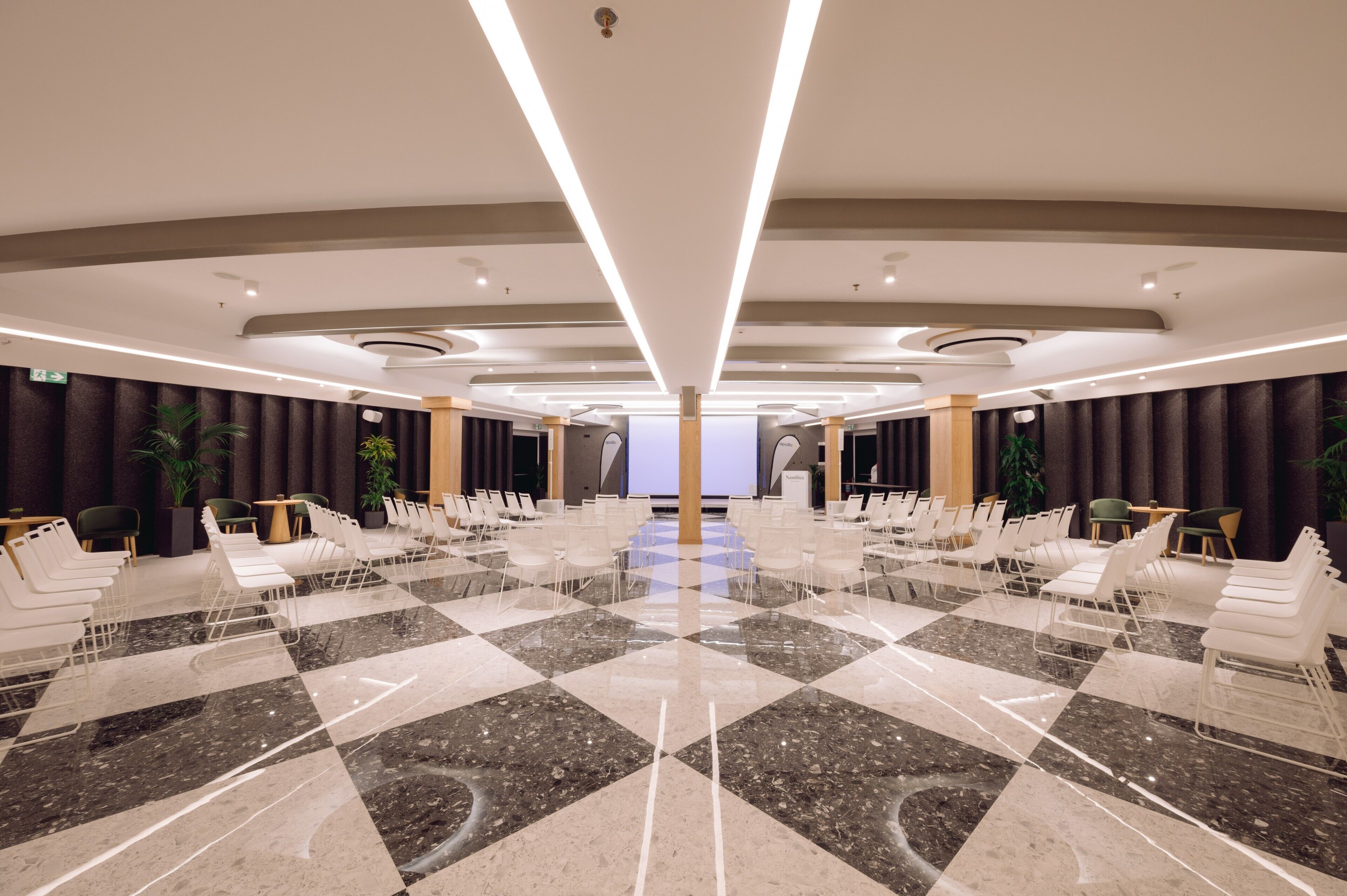 A spacious conference room with a checkered marble floor, white chairs, and a large screen at the front.