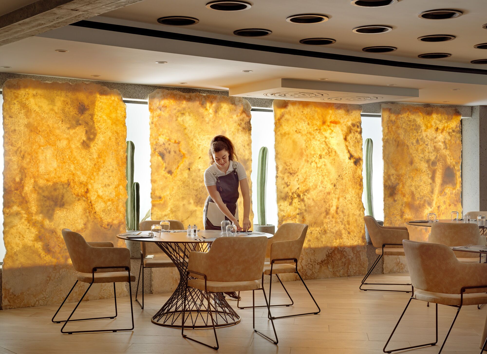 Un comedor moderno con una mesa redonda central rodeada de sillas, un camarero atendiendo a los comensales y una pared con textura al fondo.
