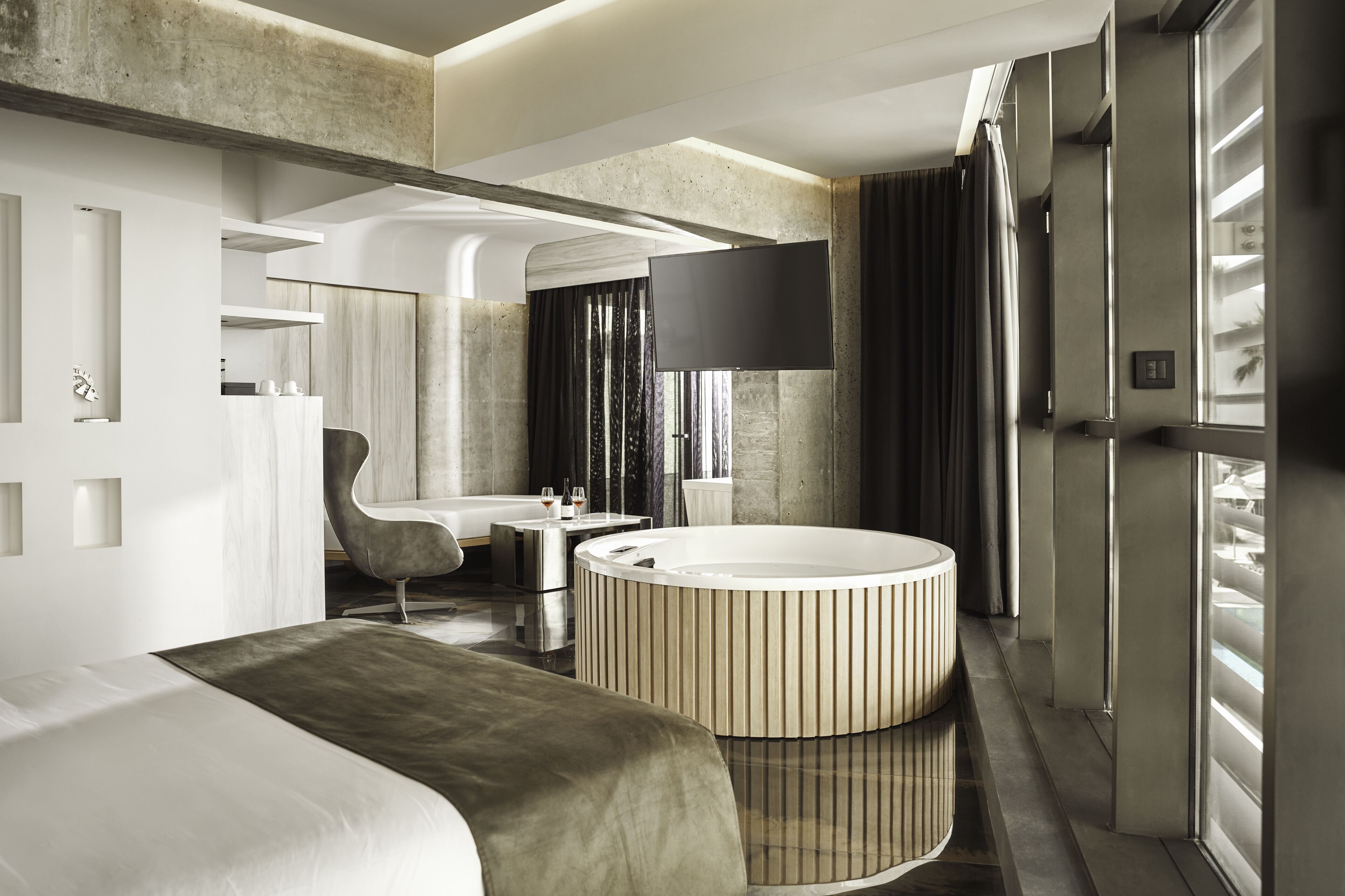 A modern hotel room with a large bed, a round bathtub, a seating area, and a flat-screen TV.
