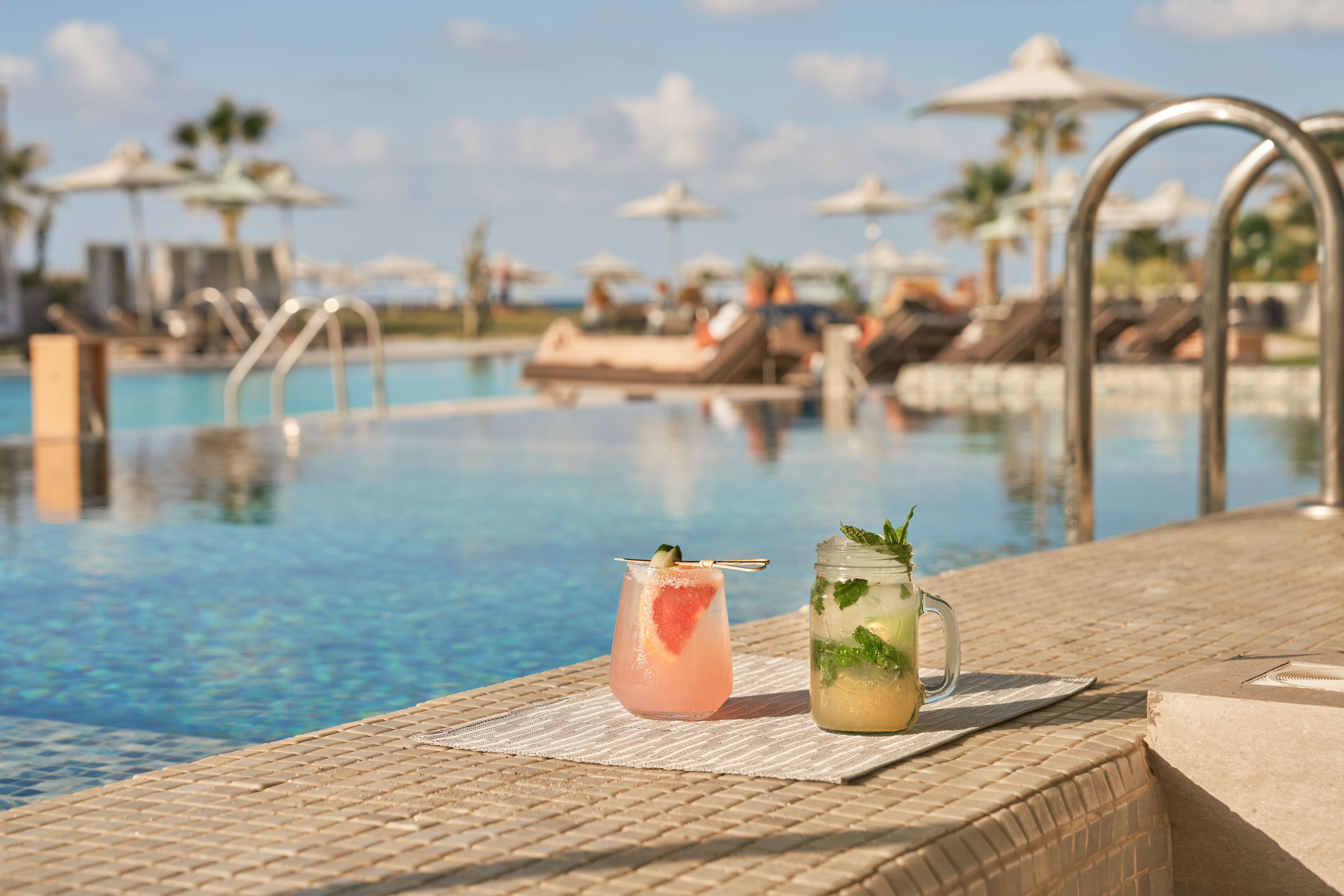 Zona de piscina con sillas reclinables y sombrillas, dos bebidas sobre una esterilla, y un cielo azul despejado al fondo.