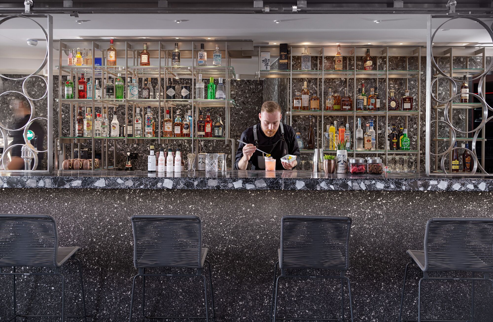 A bar area with a bartender preparing a drink, shelves of liquor bottles, and a marble countertop.