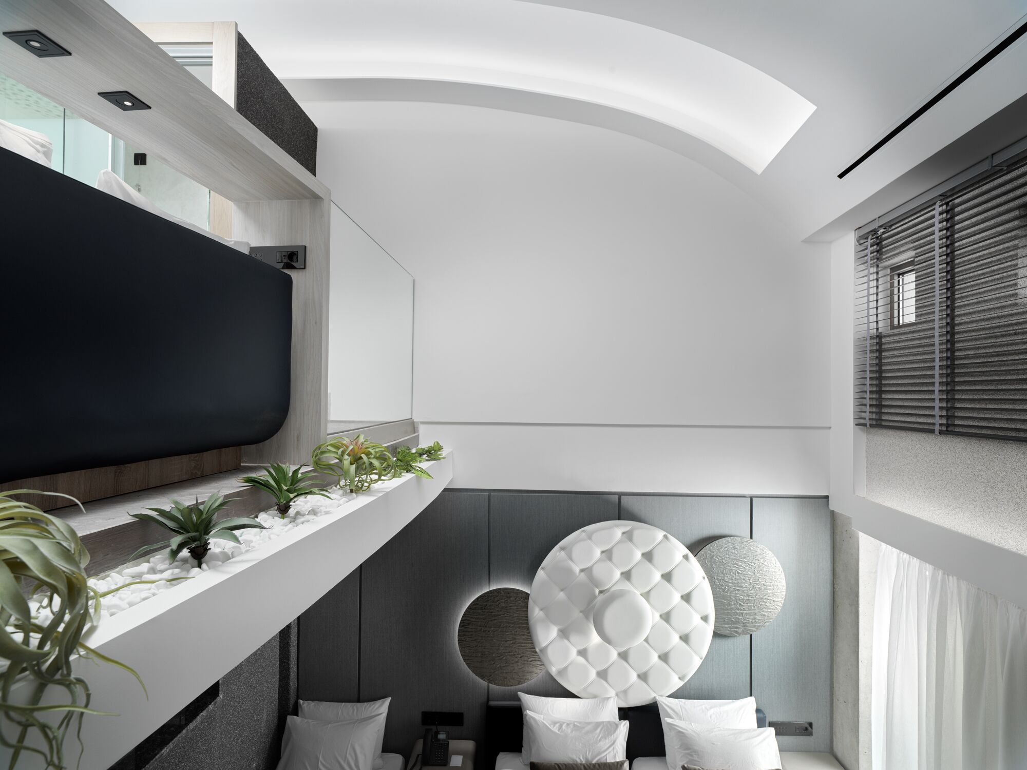 A modern hotel room with a large flat-screen TV, a bed with white bedding, a round decorative wall piece, and a plant on the windowsill.