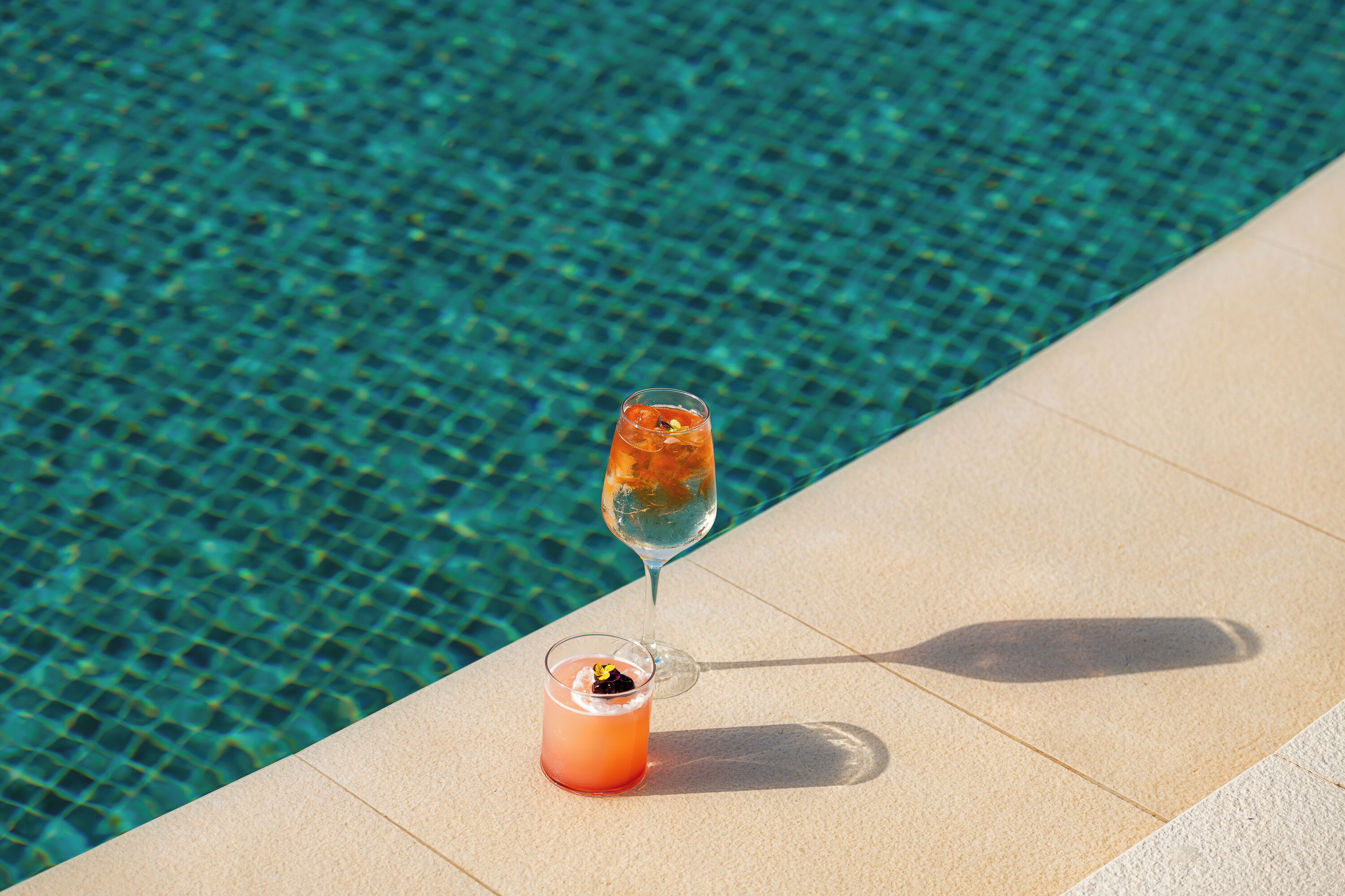 A glass of cocktail and a glass of water by a poolside.
